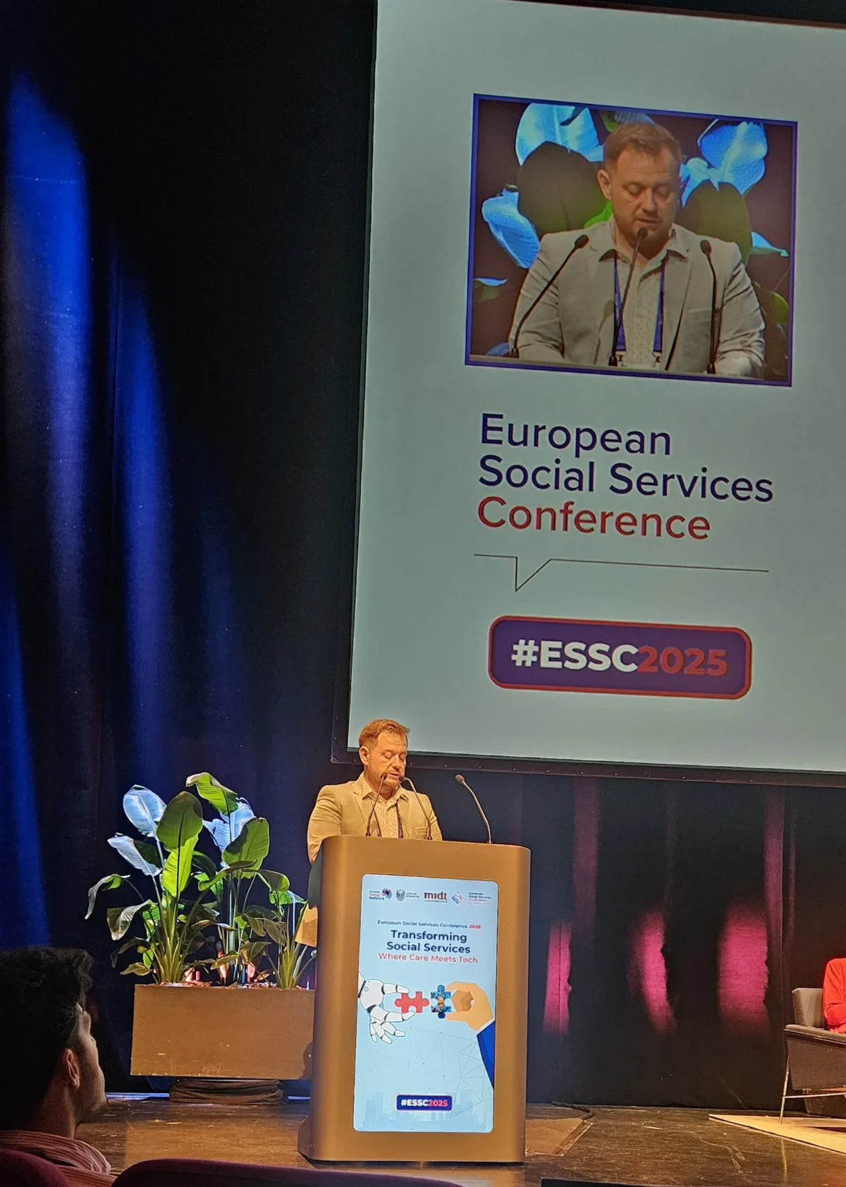 Predstavnik centra med govorom na odru konference ESSC 2025, ob njem velika projekcija s fotografijo govorca in logotipom dogodka.