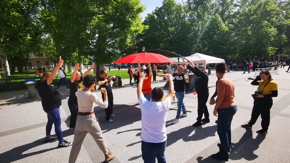 Skupina ljudi z dvignjenimi rokami sodeluje v gibalni aktivnosti na prostem.