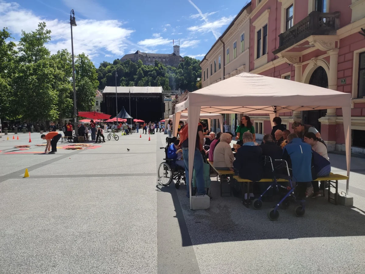 Skupina ljudi pod šotorom ustvarja in se druži ob mizi.