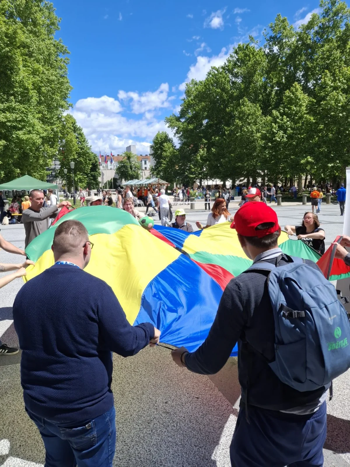 Skupina ljudi sodeluje v igri z velikim pisanim padalom na prostem.