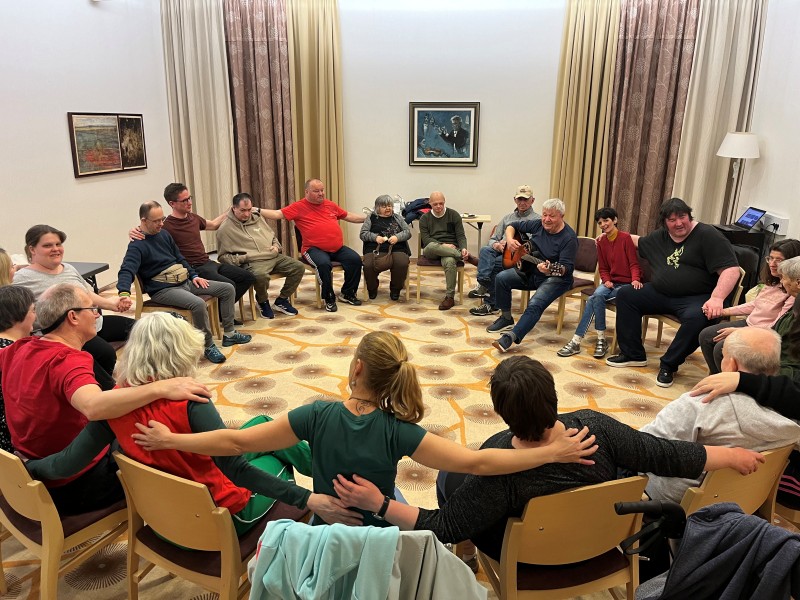 Varovanci VDC Tončke Hočevar in zaposleni sedijo na stolih v krogu, se držijo za rame in pojejo