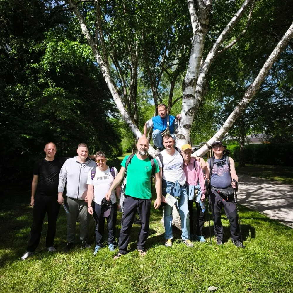Skupina udeležencev stoji na travi ob drevesu med pohodom Pot ob žici in pozira za skupinsko fotografijo.