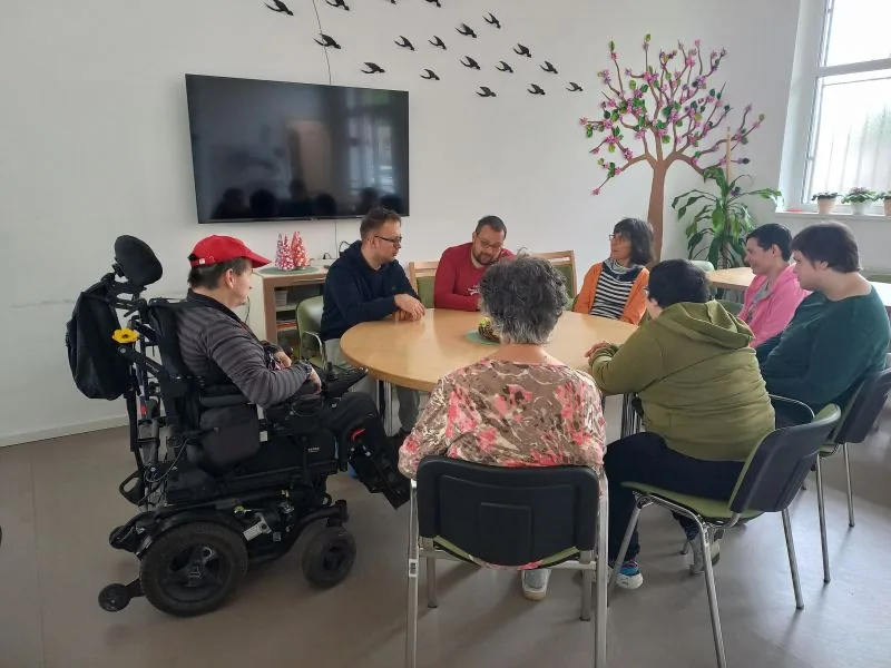 Prostovoljka Maja in uporabniki sedijo za mizo in se pogovarjajo.