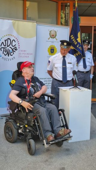 Uporabnik Boris na električnem vozičku ima nagovor na dogodku pred VDC Tončke Hočevar, v ozadju stojijo uniformirani predstavniki in zastave.