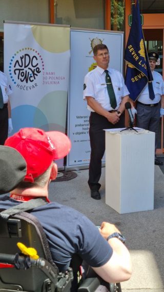 Uniformirana oseba stoji ob govorniškem pultu pred vhodom VDC Tončke Hočevar, v ospredju je udeleženec na električnem vozičku, v ozadju so zastave in predstavitveni pano.