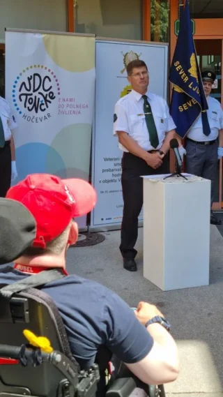 Uniformirana oseba stoji ob govorniškem pultu pred vhodom VDC Tončke Hočevar, v ospredju je udeleženec na električnem vozičku, v ozadju so zastave in predstavitveni pano.
