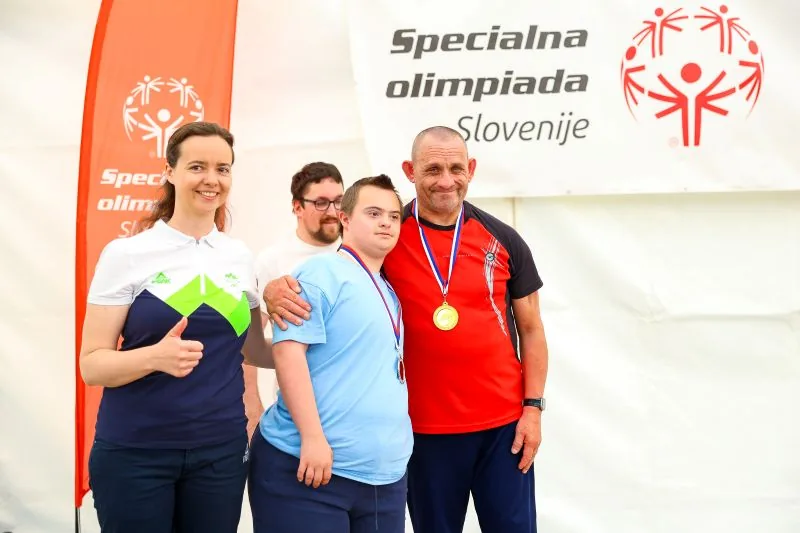Dva tekmovalca z medaljami okoli vratu objeta, zraven oseba, ki gleda v kamero in drži palec gor.