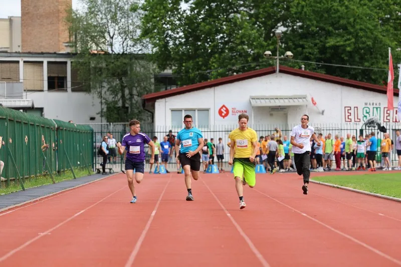 Štirje tekmovalci vzporedno tečejo na stadionu, zadaj nekaj publike in drugi tekmovalci.