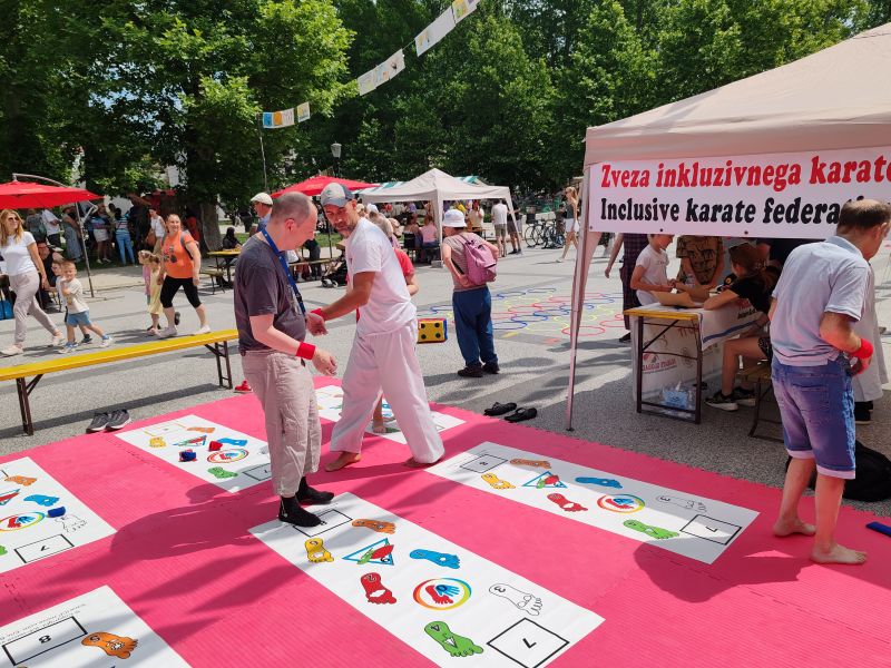 Na Kongresnem trgu poteka festivalska aktivnost na rožnati blazini s talno igro. Dve odrasli osebi izvajata vaje na označenih poljih z ilustracijami. Ob strani je bela stojnica z napisom »Zveza inkluzivnega karateja«. V ozadju so stojnice, obiskovalci in drevesa.