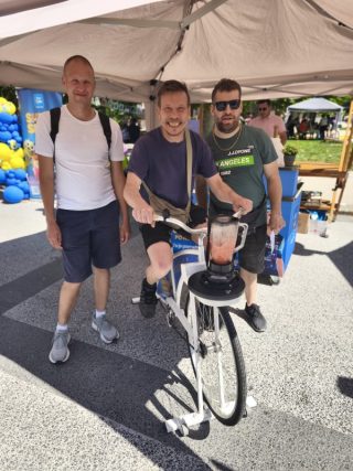 Tri odrasle osebe stojijo pod stojnico. Ena oseba sedi na posebnem kolesu, ki poganja mešalnik z napitkom. V ospredju je kolo z nameščenim mešalnikom, v ozadju pa festivalski prostor z dodatnimi stojnicami.