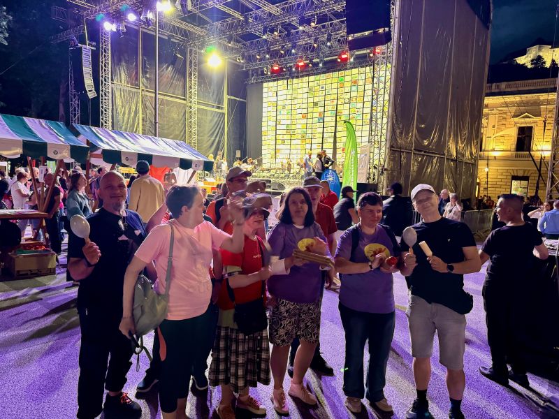 Nekaj naših uporabnikov stoji pred osvetljenim odrom in drži glasbene rekvizite. Oder je razsvetljen z barvnimi lučmi, okoli pa so obiskovalci festivala in stojnice v večernem ambientu.