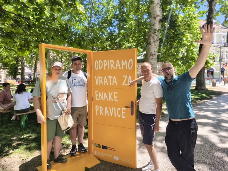 Na prostem stoji velika rumena umetniška instalacija v obliki odprtih vrat z napisom »ODPIRAMO VRATA ZA ENAKE PRAVICE«. Ob instalaciji stojijo štiri odrasle osebe, v ozadju pa so drevesa in mestni park.