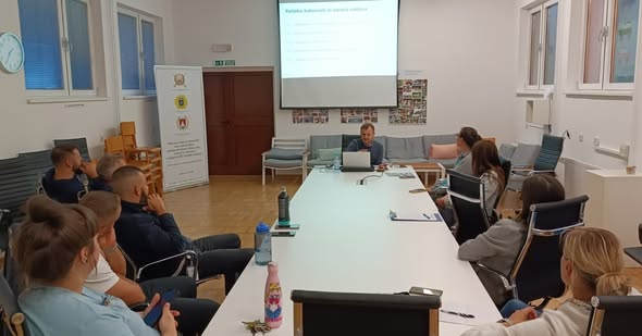 Skupina zaposlenih sedi za veliko mizo v sejni sobi in spremlja predavanje s predstavitvijo na projekcijskem platnu.