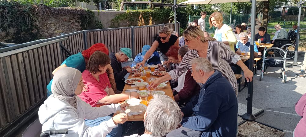 Udeleženci piknika VDC Tončke Hočevar sedijo za skupno mizo na prostem, zaposleni pa jim postrežejo s hrano in pijačo.