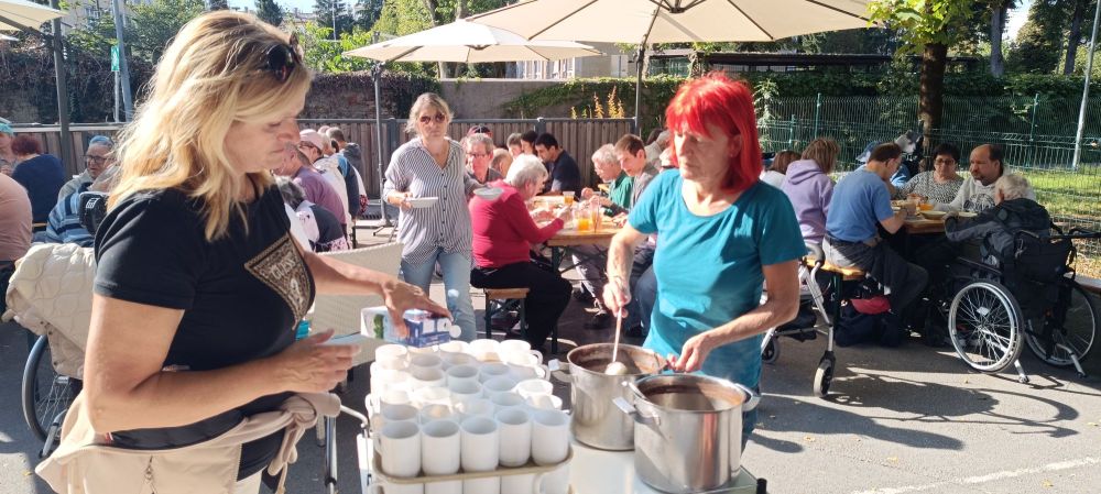 Na pikniku VDC Tončke Hočevar dve osebi pripravljata in delita tople napitke, medtem ko udeleženci sedijo za mizami na prostem.