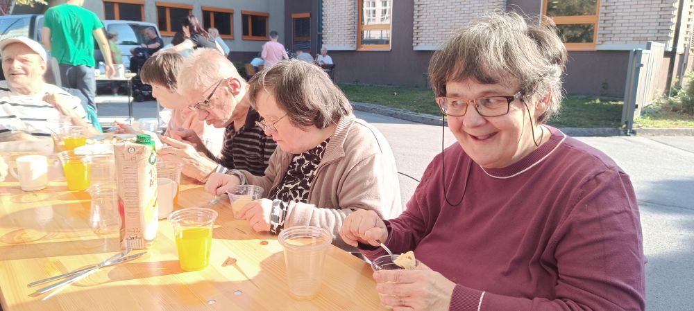 Udeleženka piknika VDC Tončke Hočevar sedi za mizo na prostem in uživa sladoled, medtem ko drugi udeleženci obedujejo v ozadju.
