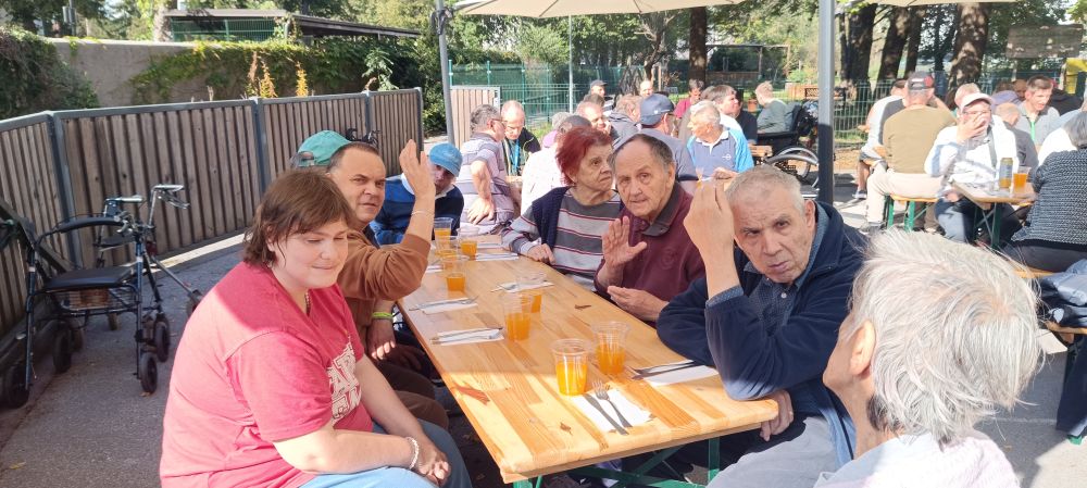 Udeleženci piknika VDC Tončke Hočevar sedijo za skupno mizo in se pogovarjajo ob osvežilnih napitkih.