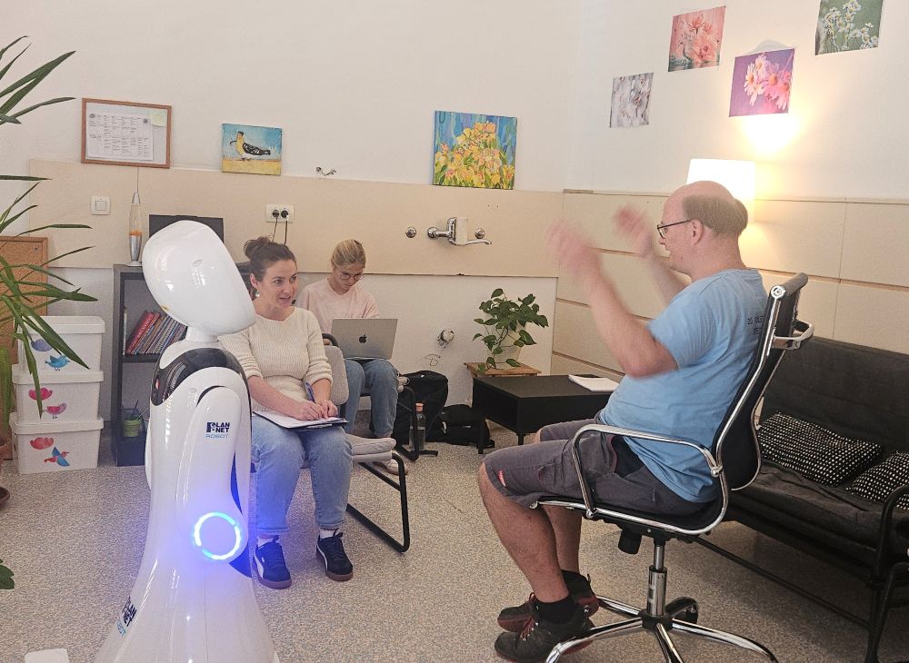 Socialni robot Timo med preizkusno interakcijo z uporabnikom VDC Tončke Hočevar, raziskovalci pa spremljajo potek kognitivnega treninga.