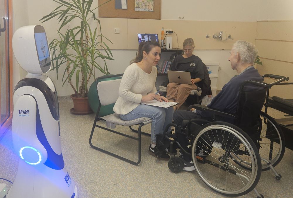 Socialni robot Timo sodeluje pri preizkusni interakciji z uporabnikom VDC Tončke Hočevar, medtem ko raziskovalci spremljajo potek aktivnosti.