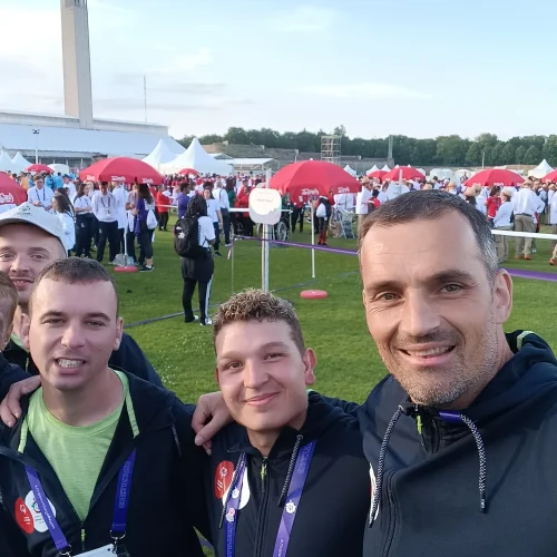 Pet nasmejanih moških z akreditacijami na prizorišču športnih iger, v ozadju šotori in obiskovalci