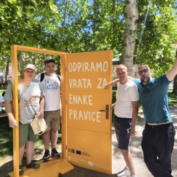 Na prostem stoji velika rumena umetniška instalacija v obliki odprtih vrat z napisom »ODPIRAMO VRATA ZA ENAKE PRAVICE«. Ob instalaciji stojijo štiri odrasle osebe, v ozadju pa so drevesa in mestni park.