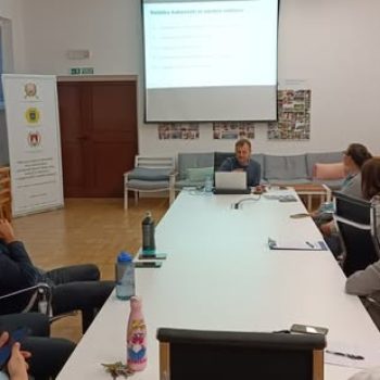 Skupina zaposlenih sedi za veliko mizo v sejni sobi in spremlja predavanje s predstavitvijo na projekcijskem platnu.